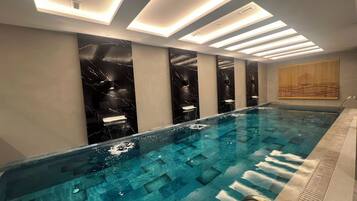 A modern indoor swimming pool with a clear water surface, a metal ladder, and a series of recessed lighting panels on the ceiling.