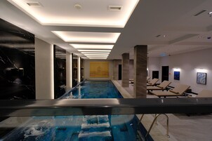 Piscina interior moderna com barreira de vidro, cadeiras de descanso e iluminação embutida.