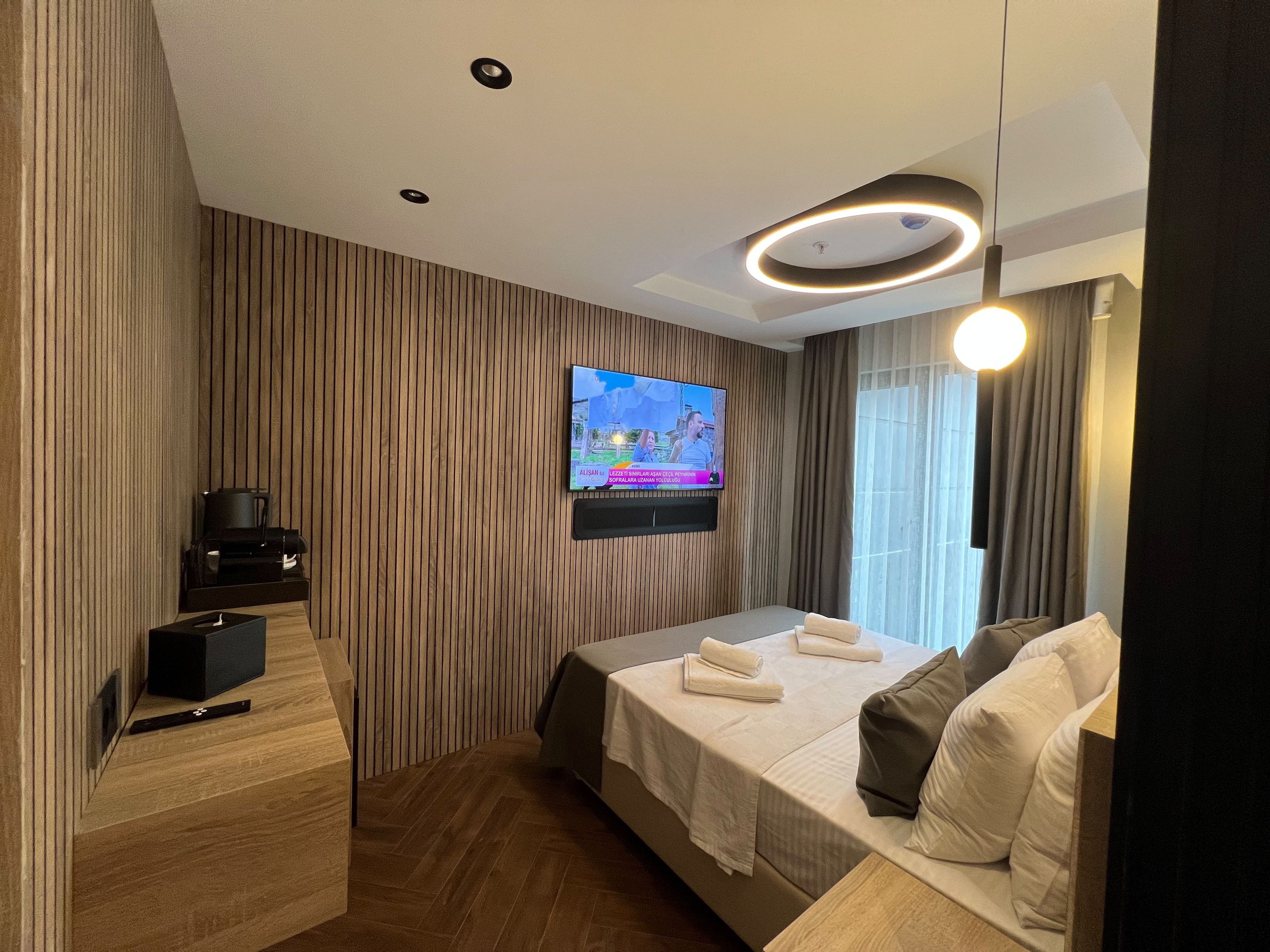 Una habitación de hotel moderna con un televisor de pantalla plana montado en la pared, una cama con toallas ordenadas, una cafetera y un panel de pared de madera.