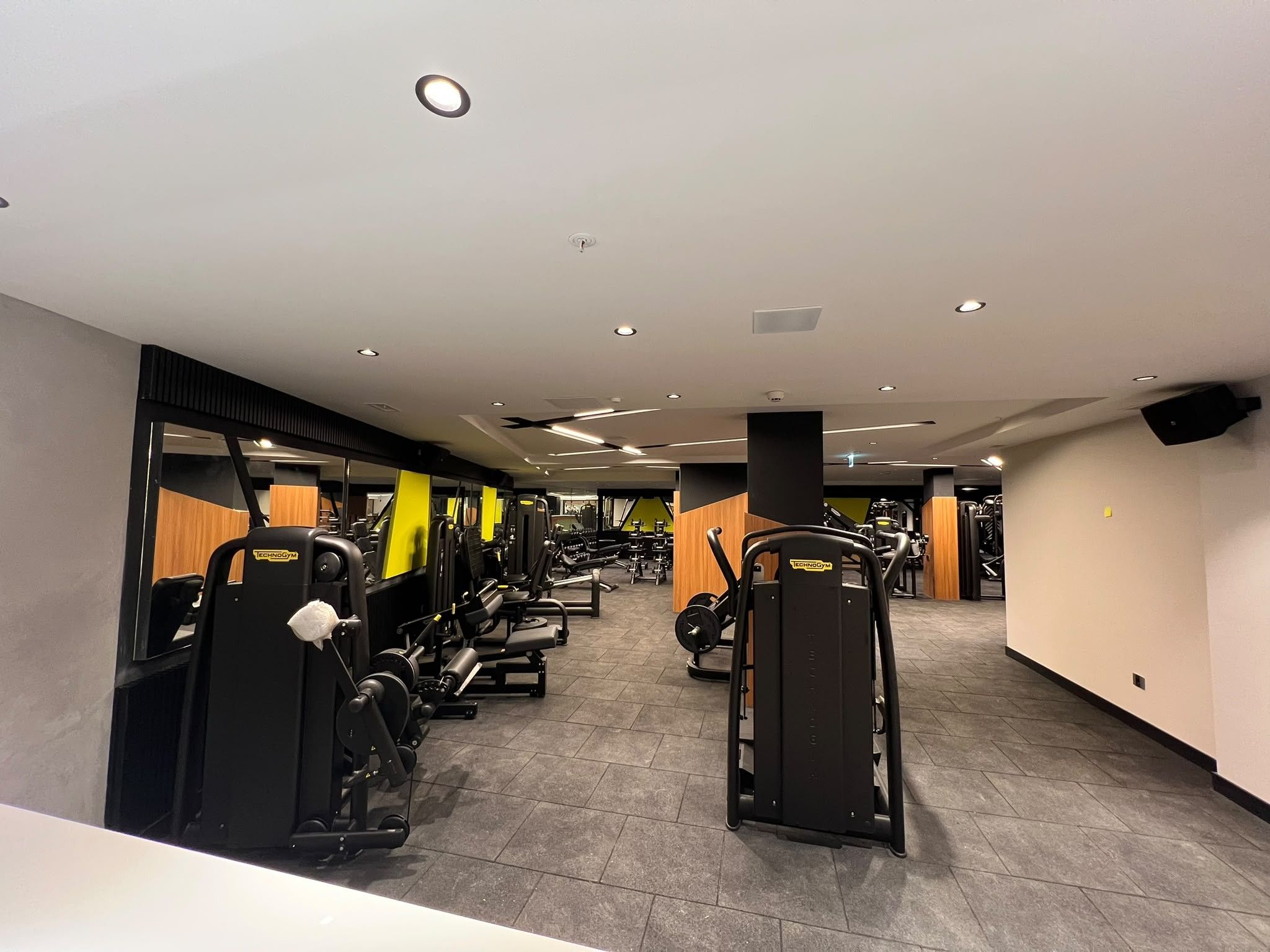 Un gimnasio con diversas máquinas de ejercicio, piso de baldosas e iluminación empotrada en el techo.