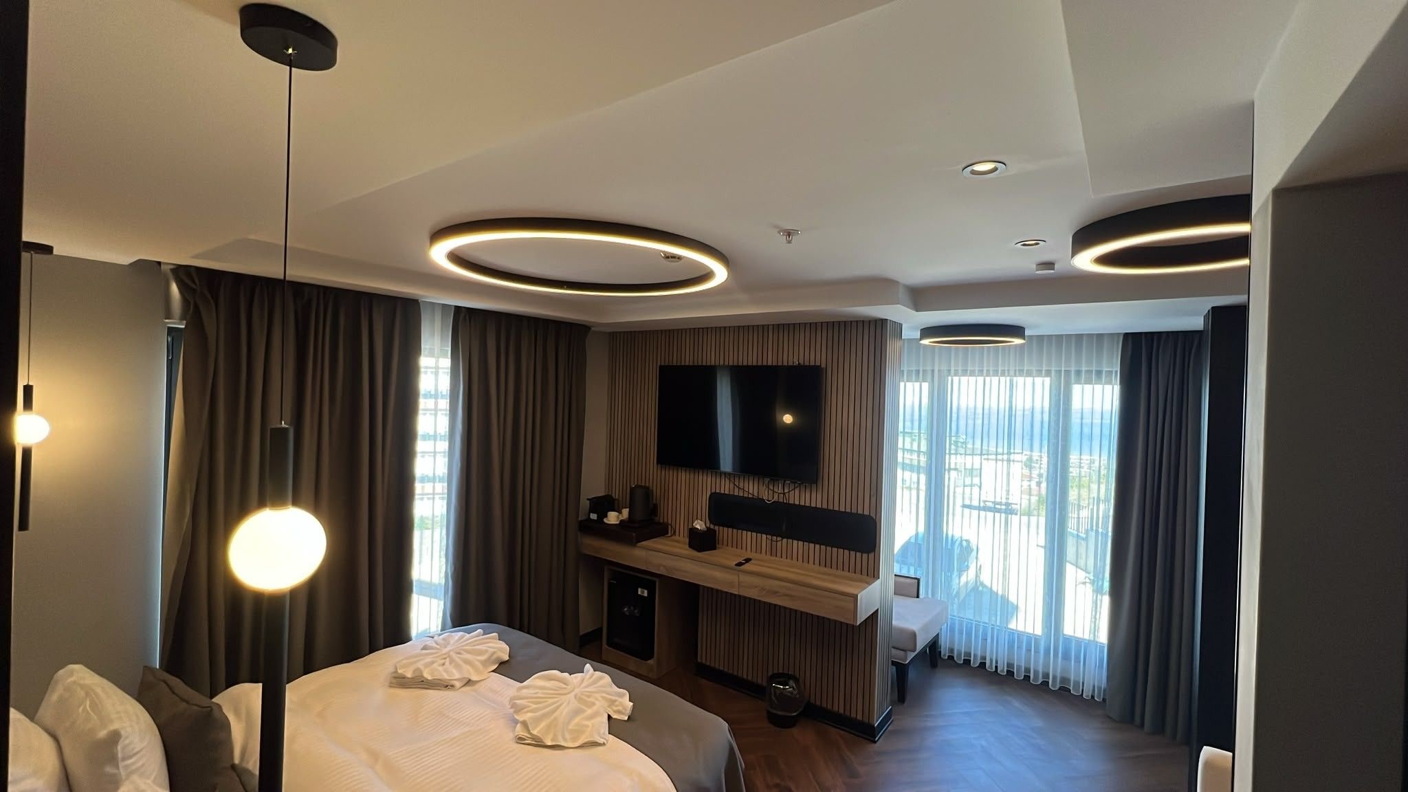 Una habitación de hotel moderna con una cama grande, un televisor de pantalla plana, un escritorio con una silla y un ventanal con cortinas.