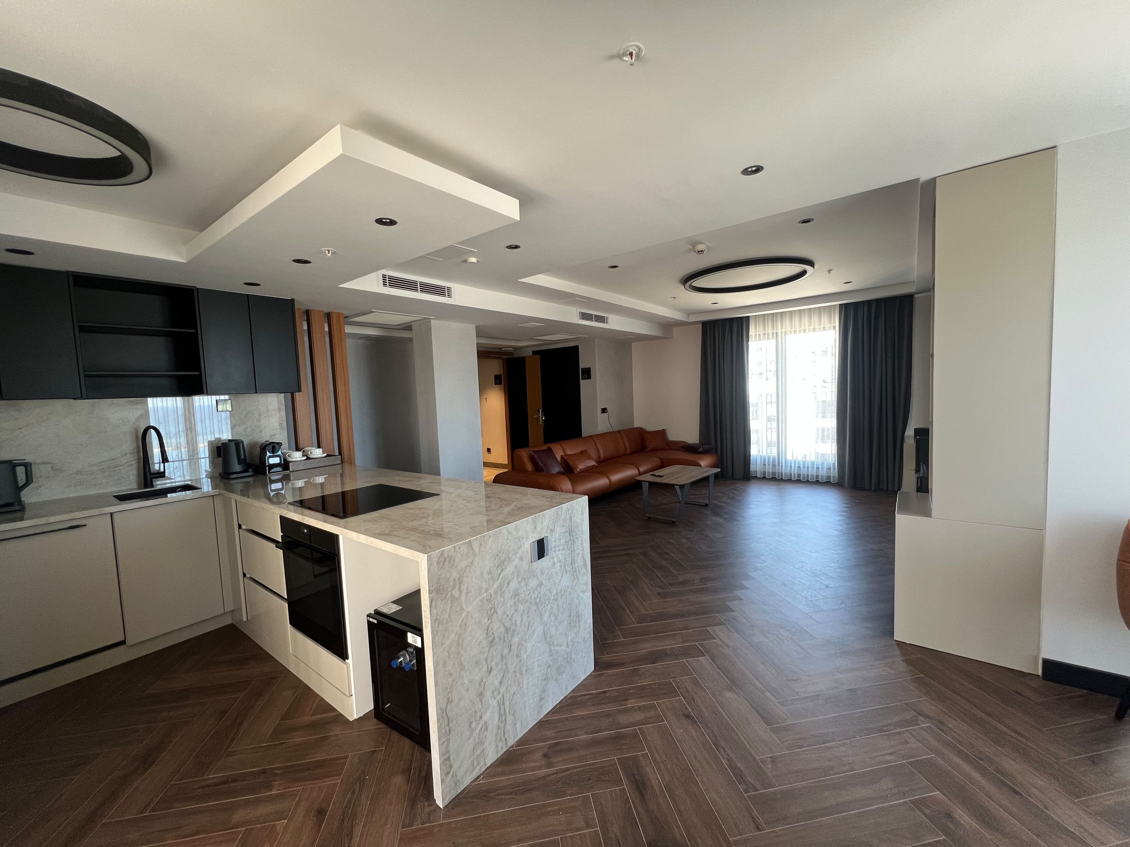 A modern kitchen with a central island, dark wood flooring, and a living area with a leather sofa.