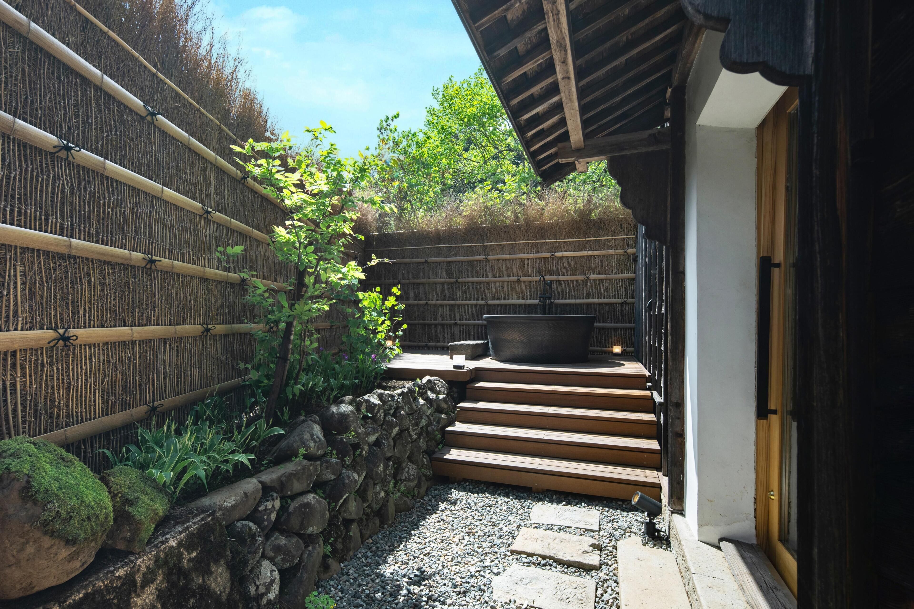 A traditional Japanese-style garden with a stone wall, bamboo fence, and a wooden staircase leading to a building entrance.