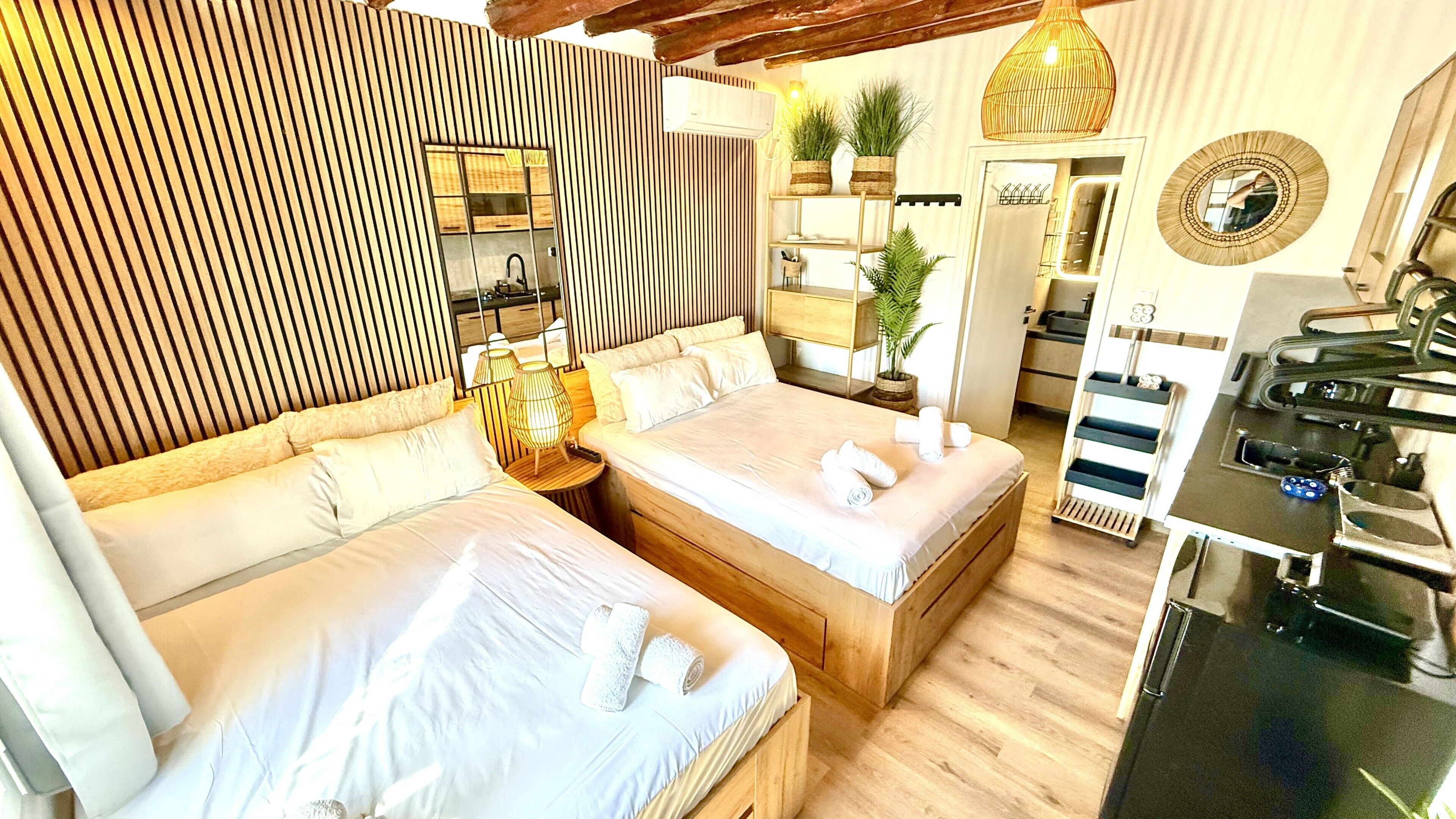 A modern hotel room with a large bed, a small desk, a mirror, and a kitchen area.