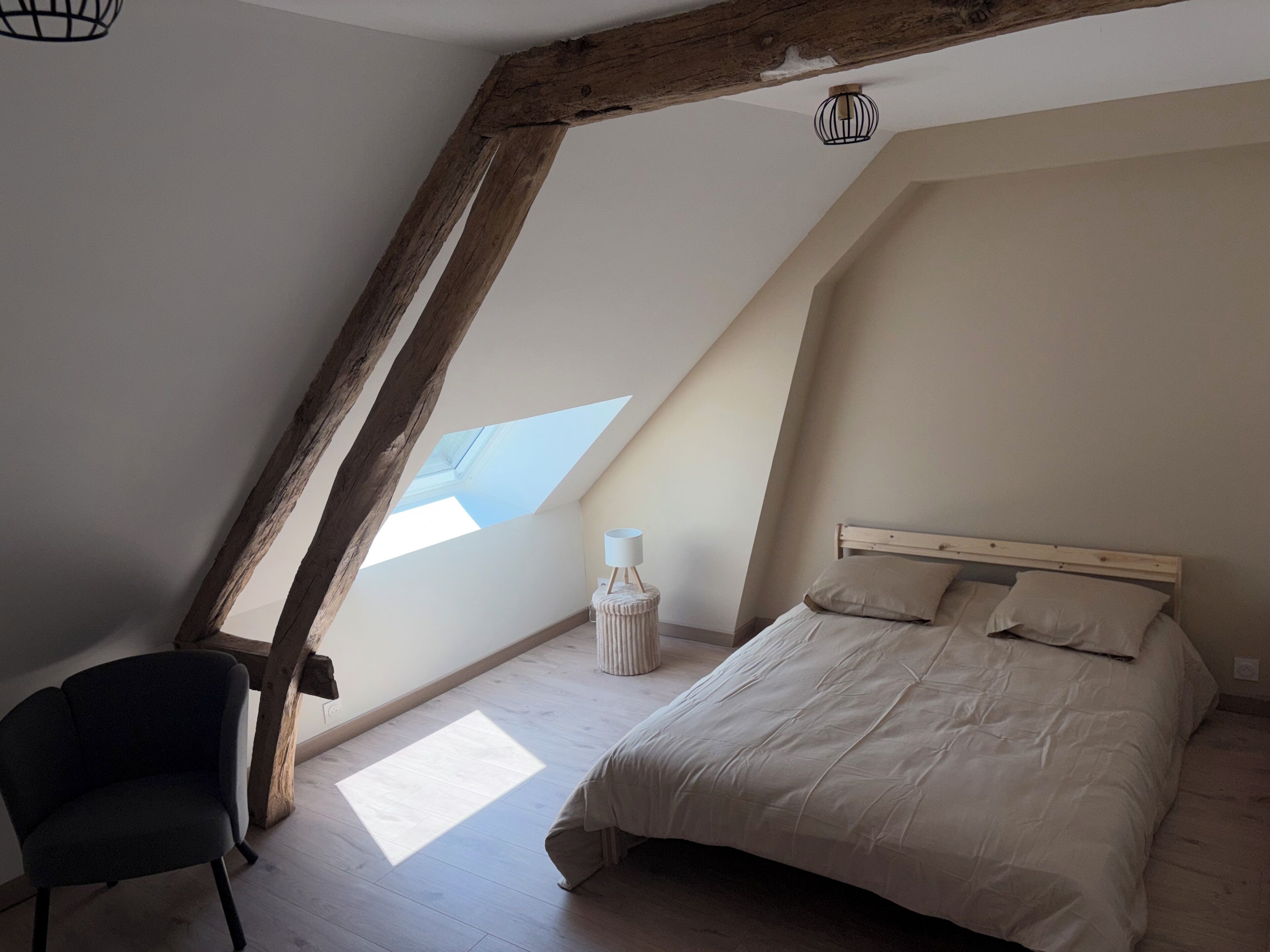A bedroom with a sloped ceiling, a skylight, a bed with pillows, a bedside lamp, and a chair.