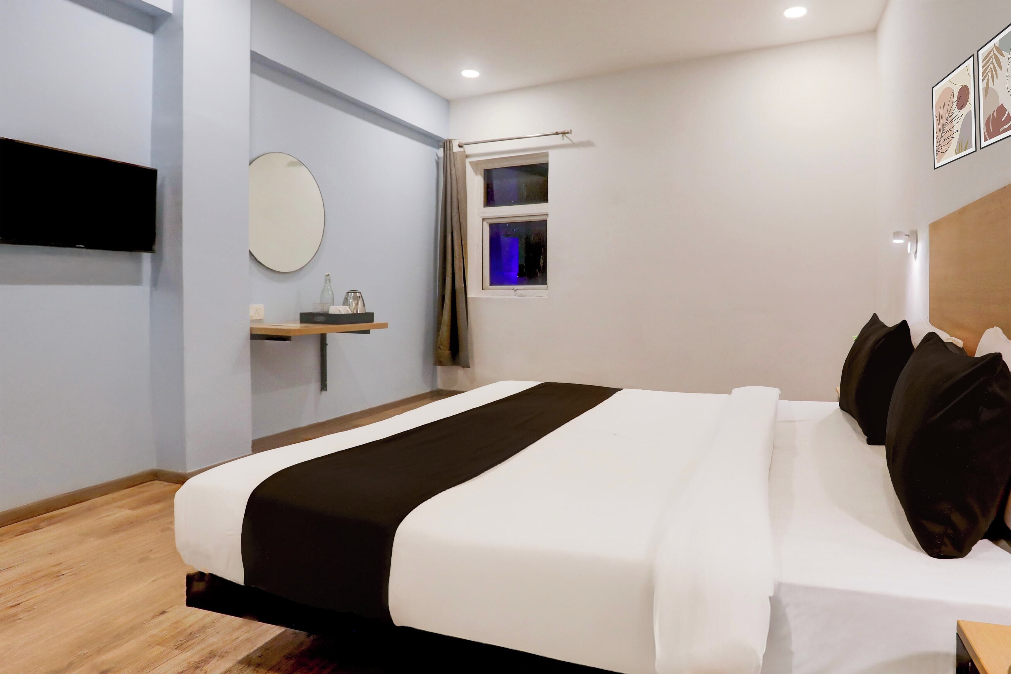A modern hotel room with a large bed, a flat-screen TV, a bedside table, a window with curtains, and a wall-mounted mirror.