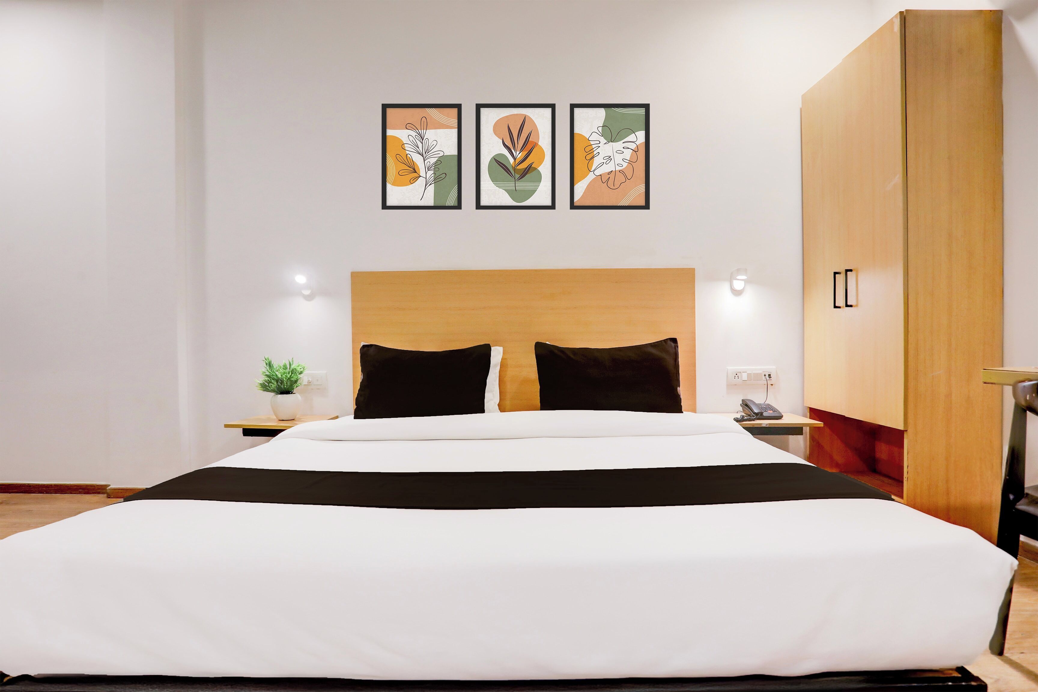 A modern hotel room with a large bed, a wooden headboard, a bedside table with a plant, and a wardrobe.