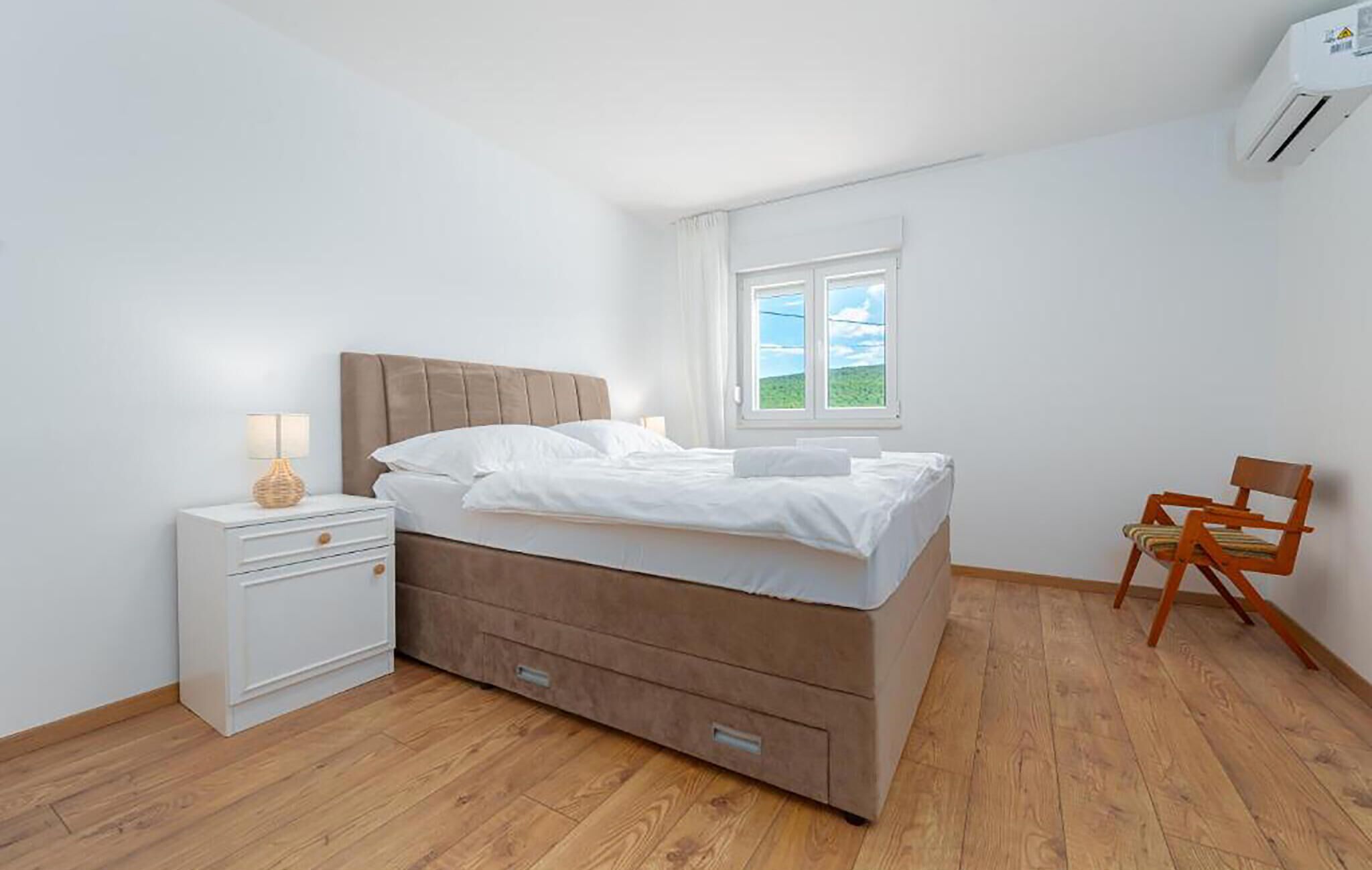 A bedroom with a bed, a nightstand, a chair, a window, and an air conditioning unit.