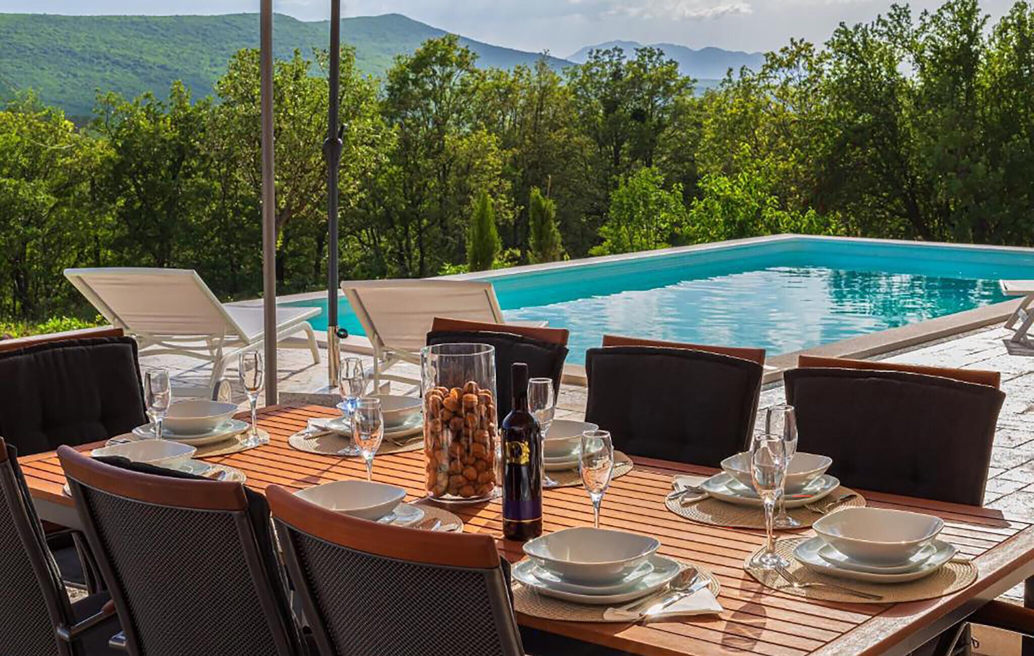 A dining table set for two with a view of a swimming pool and mountains in the background.
