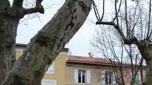 A building with a yellow facade, white shutters, and a balcony. There are two cars parked in front. - The perfect getaway on the square in Saint-Chinian, home to its famous wine (Saint-Chinian)
