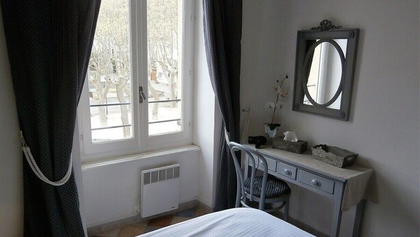 A bedroom with a bed, a desk, a chair, a radiator, and a window with curtains. - The perfect getaway on the square in Saint-Chinian, home to its famous wine (Saint-Chinian)
