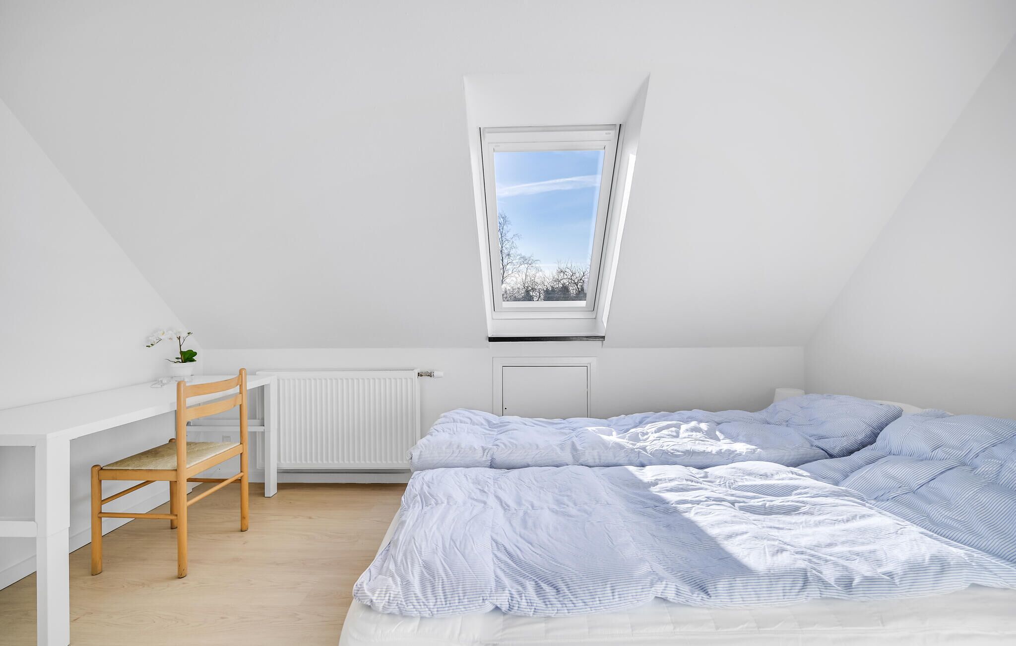 A bedroom with a sloped ceiling, a skylight, a radiator, a wooden chair, and a bed with striped bedding.