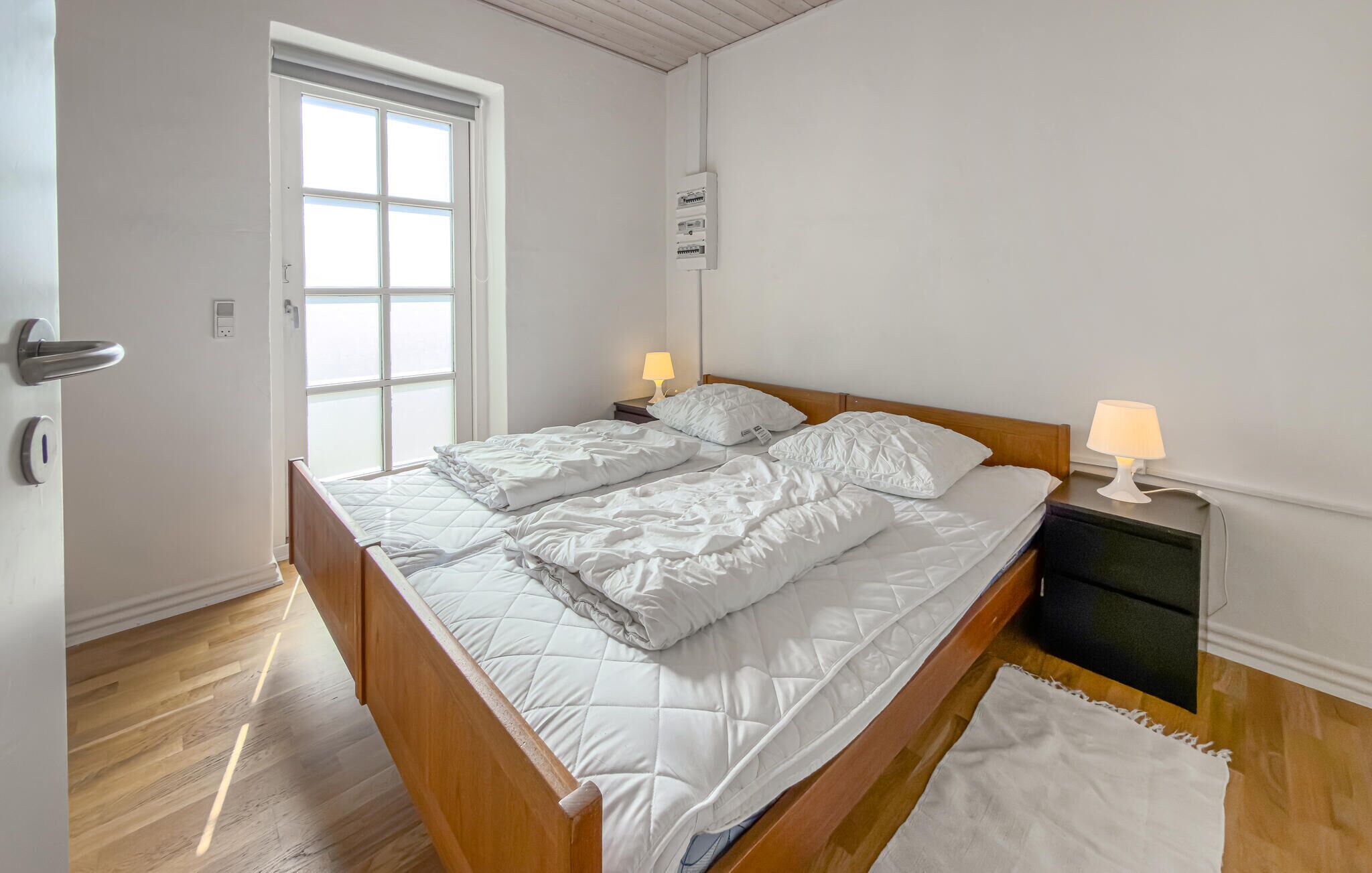 A bedroom with a wooden bed, two bedside lamps, a window, and a wall-mounted electrical box.