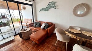 Ein Wohnzimmer mit einem braunen Ledersofa, einem runden Couchtisch und einem weißen Sessel. Ein großes Fenster bietet Blick ins Freie.