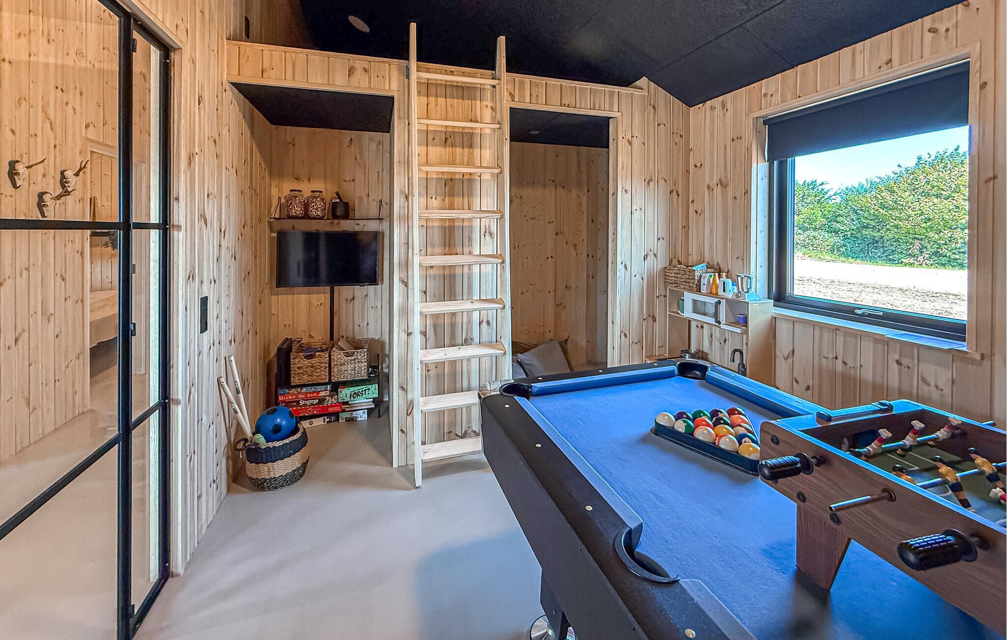 A pool table with a foosball table, a ladder, and shelves with items in a room with wooden walls and a window.