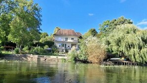 Een huis met een vijver ervoor, omringd door bomen en een heldere lucht.