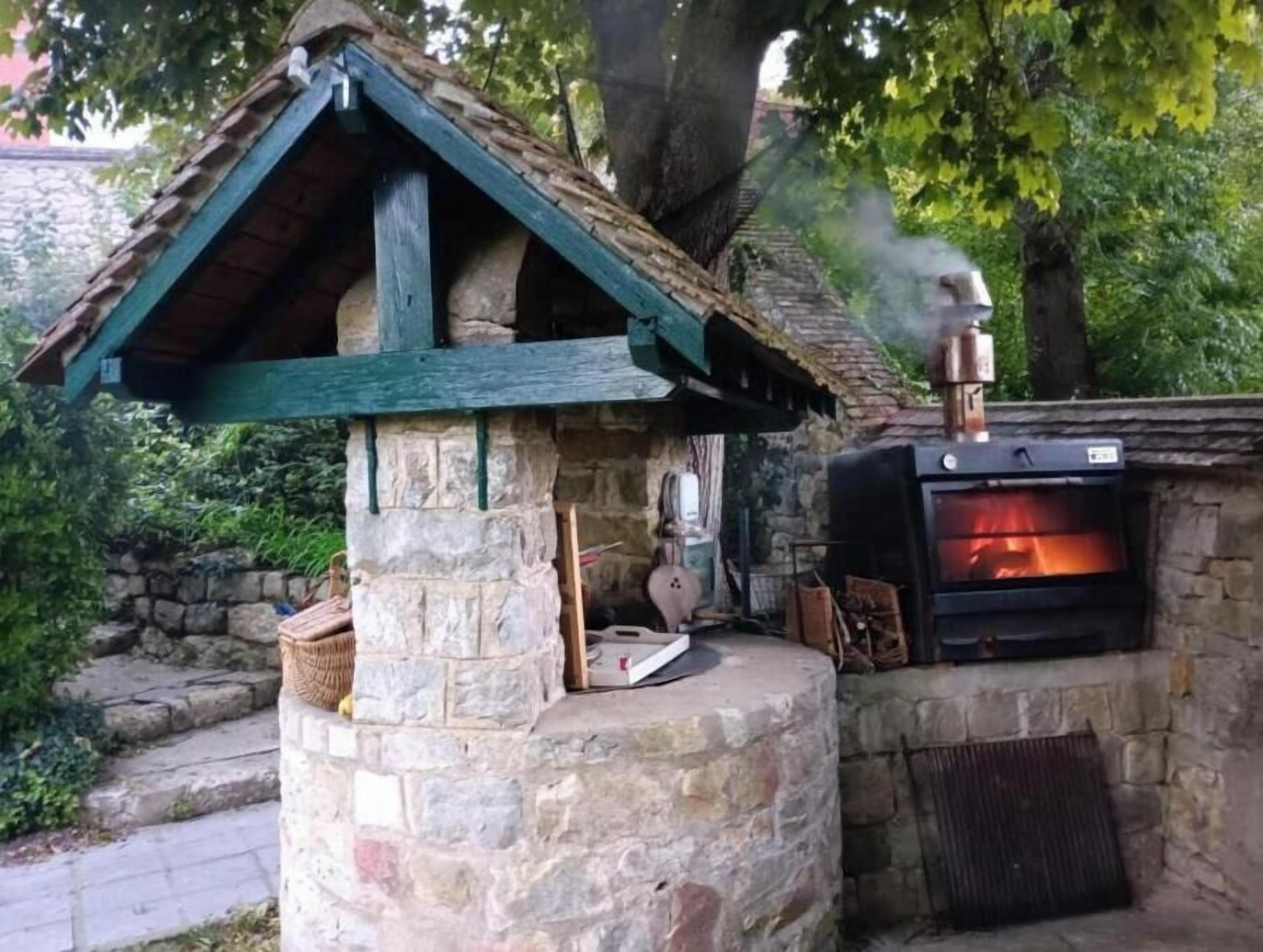 Un four extérieur en pierre, doté d’une cheminée qui dégage de la fumée, est entouré de verdure.