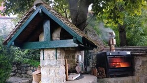 Forno de pedra ao ar livre com chaminé soltando fumaça, rodeado por vegetação.