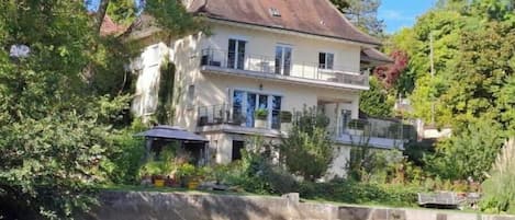 A two-story house with a balcony, situated beside a body of water, surrounded by greenery.