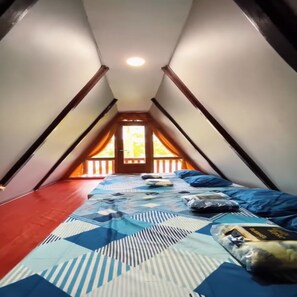Ein schmales Schlafzimmer im Dachgeschoss mit schrägen Wänden, einem Bett mit gemusterten Textilien und Blick durch eine Tür nach draußen.