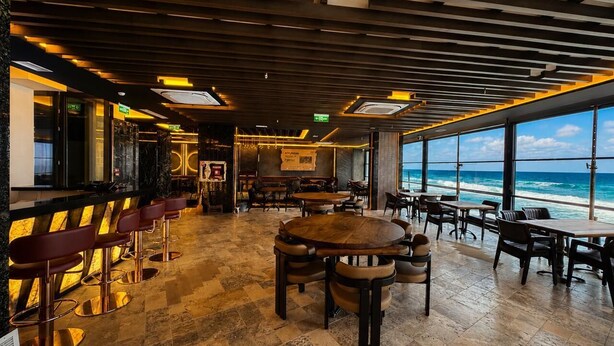 Restaurante con vista al océano, con mesas y sillas de madera, una zona de bar con taburetes y un diseño de techo moderno.
