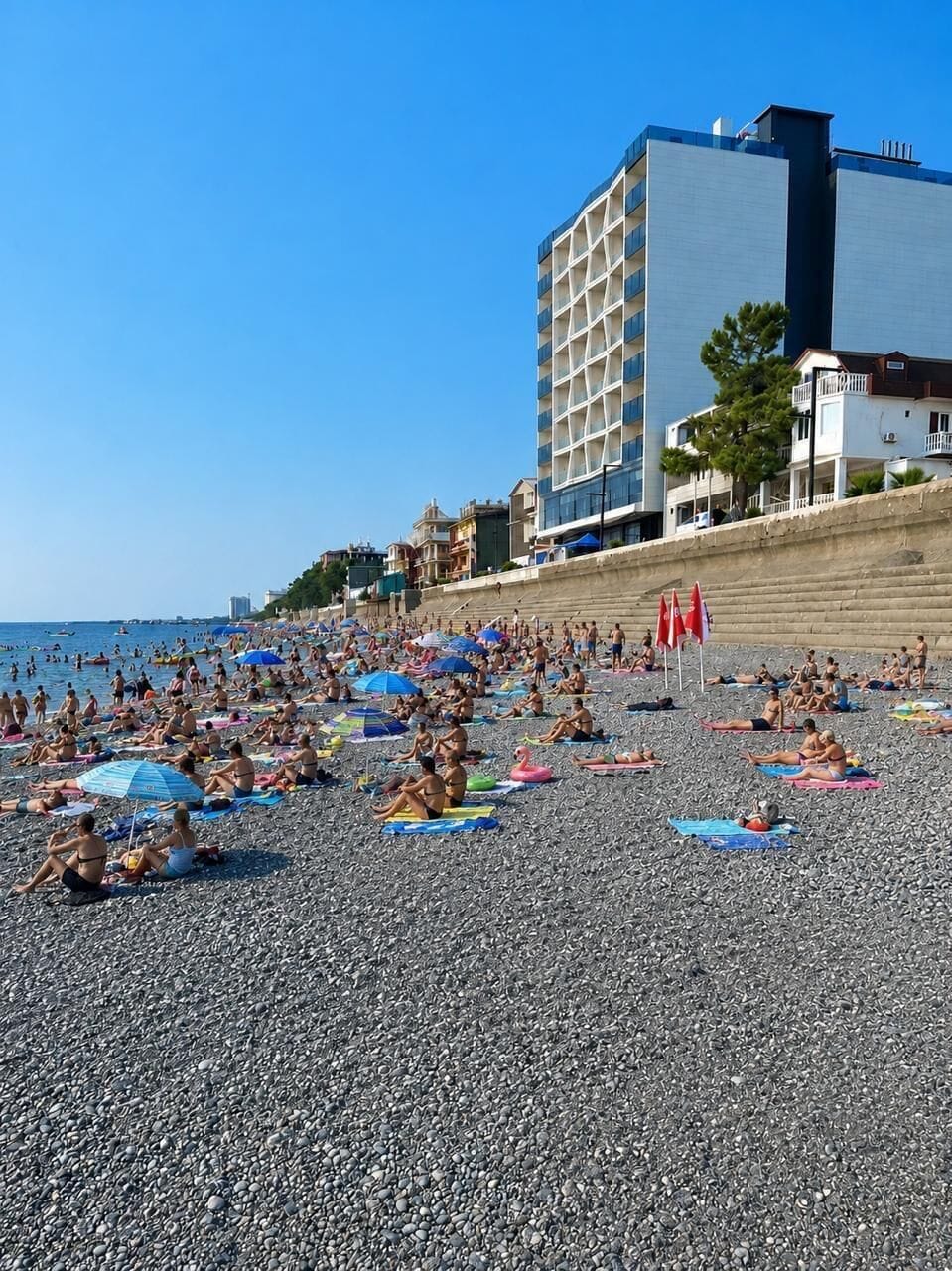 A pebble beach with people sunbathing and swimming, a tall building, and a clear blue sky.