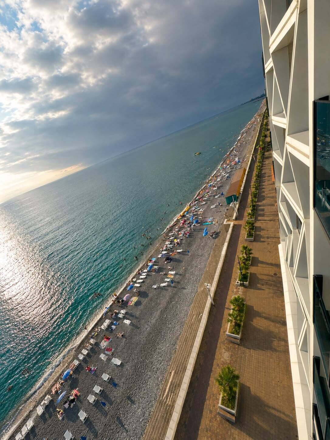 Una playa con sombrillas y tumbonas, un paseo marítimo con macetas y un edificio de diseño moderno.