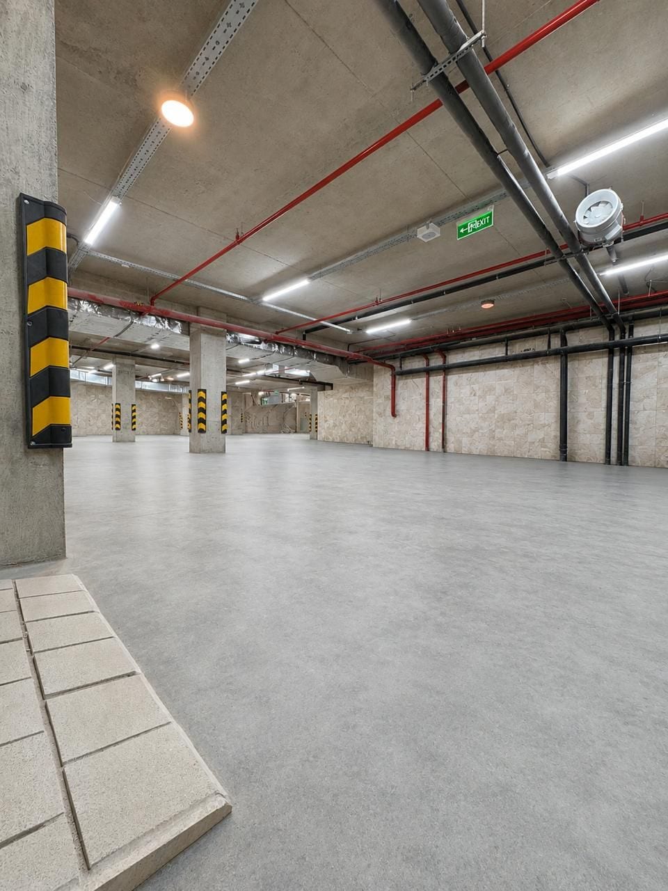 An empty parking garage with concrete pillars, red pipes, and an exit sign.