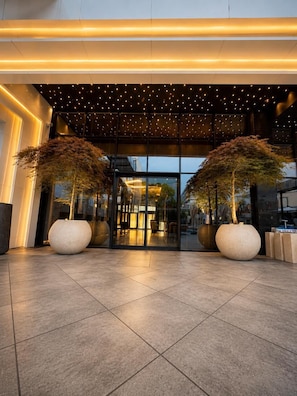 A modern building entrance with a glass facade, large potted trees, and a tiled floor. - The New Jai Son (Kobuleti)