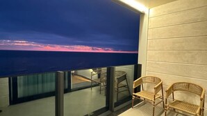Un balcón con vista al océano al atardecer, barandilla de cristal y dos sillas de mimbre.