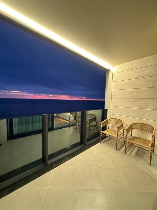 A balcony with a view of the ocean at sunset, a glass railing, and two wicker chairs.