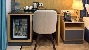 A hotel room with a minibar, a desk with a kettle, a chair, a bedside table with a lamp, and a bed.