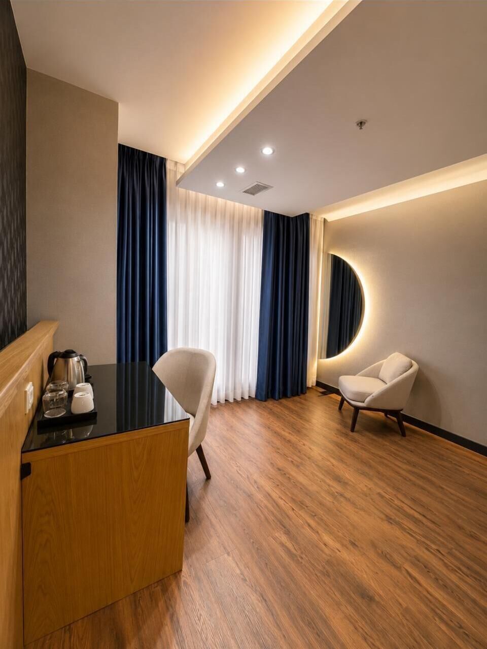 Una habitación de hotel moderna con piso de madera, un escritorio con tetera, dos sillas y un amplio ventanal con cortinas.