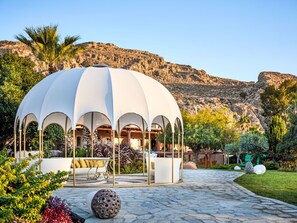 Um gazebo branco com arcos, um caminho de pedra e montanhas ao fundo.