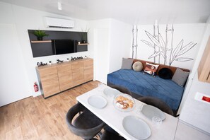 Ein gemütlicher Raum mit einem Bett, einem Esstisch mit Stühlen, einem Fernseher und einem Wandregal mit Pflanzen.