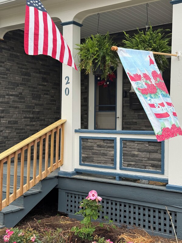 A house with a stone facade, a wooden staircase, and a porch with a hanging American flag and a banner.