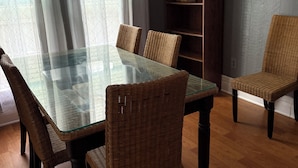 Un coin repas avec une table à plateau de verre, des chaises en osier, une bibliothèque en bois et une vue sur l’extérieur à travers une fenêtre ornée de rideaux.