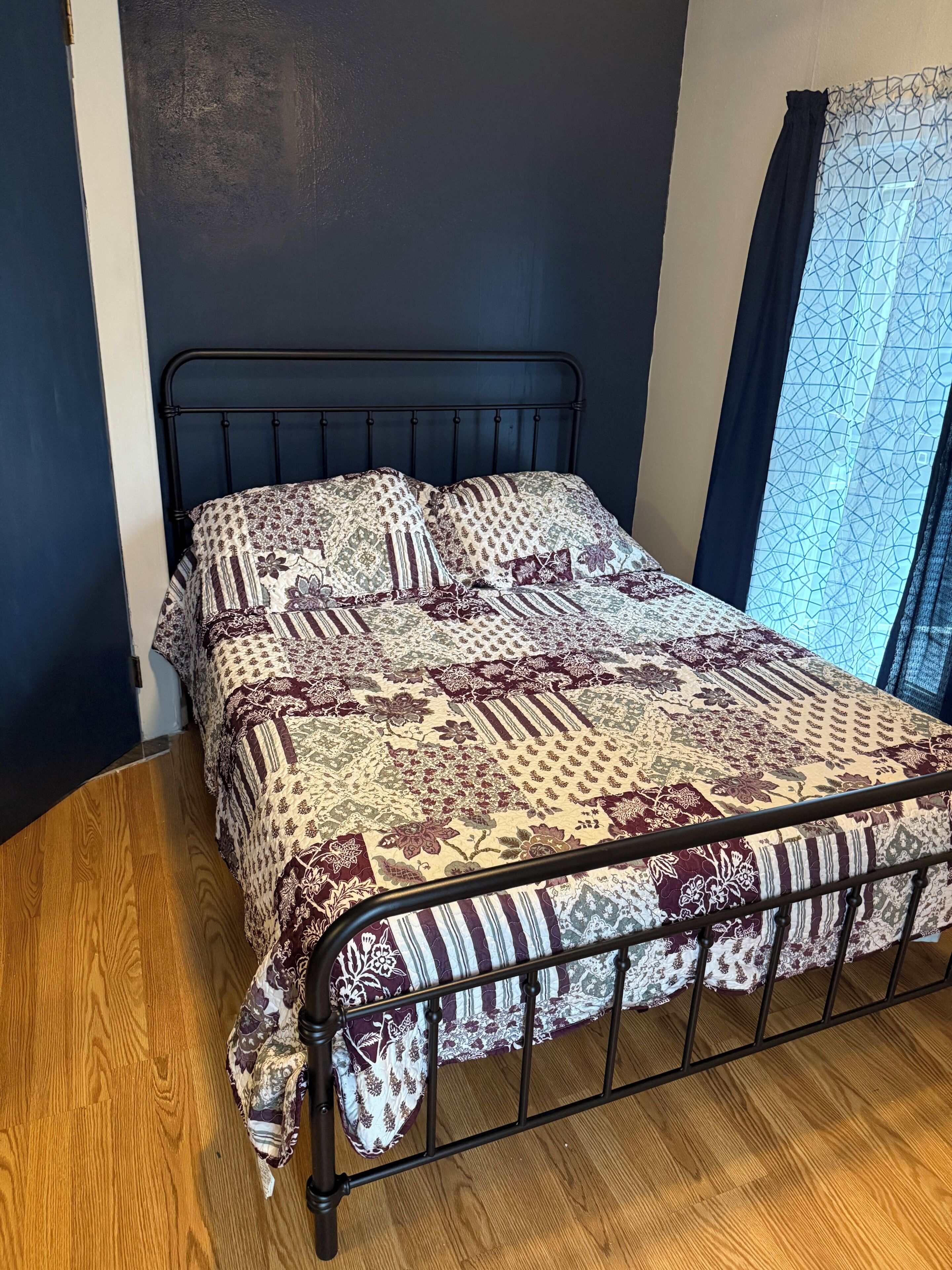 Una cama con una colcha estampada, un cabecero oscuro y una ventana con cortina geométrica.