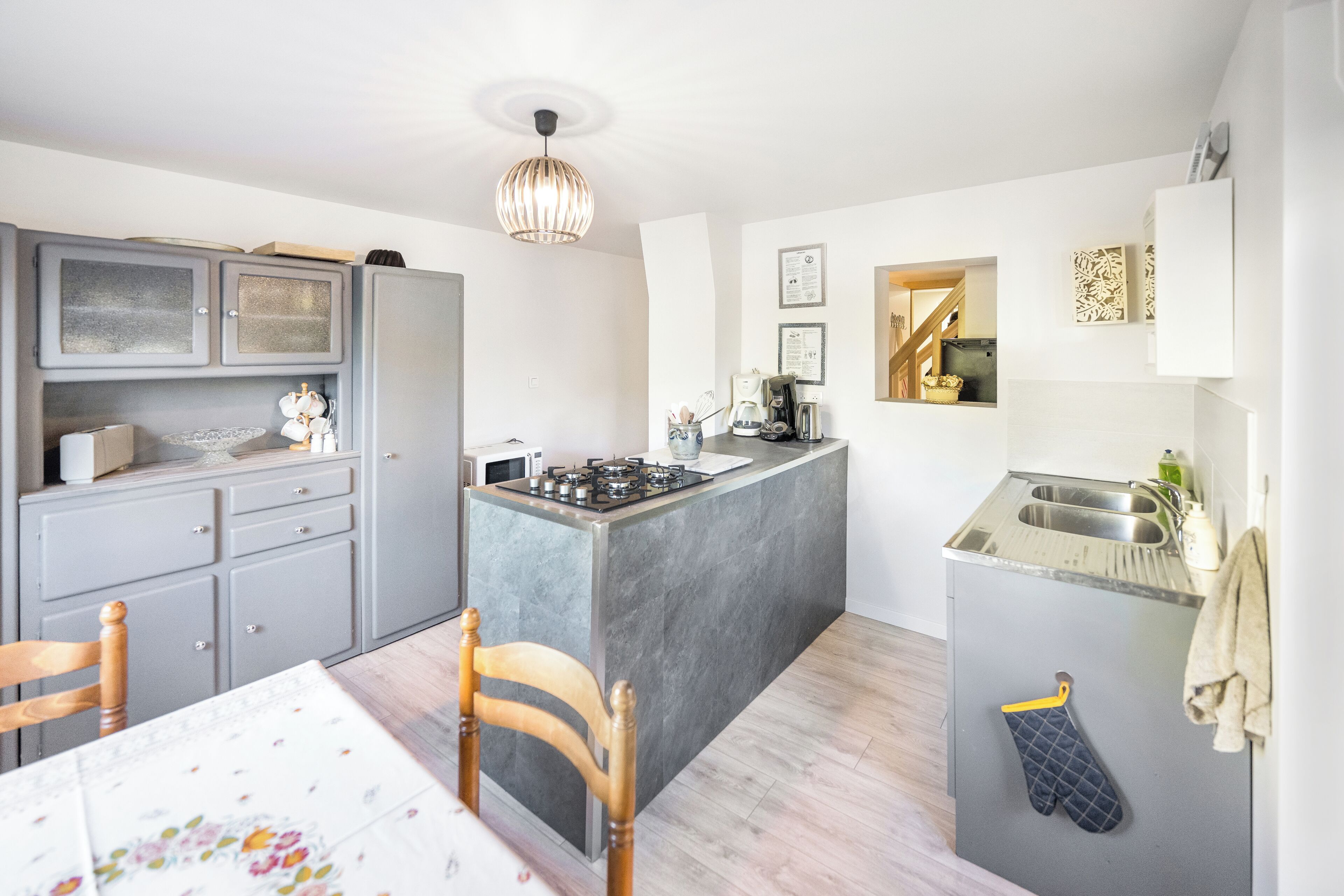A compact kitchen with a central island, grey cabinetry, a dining table with chairs, and a hanging oven mitt.