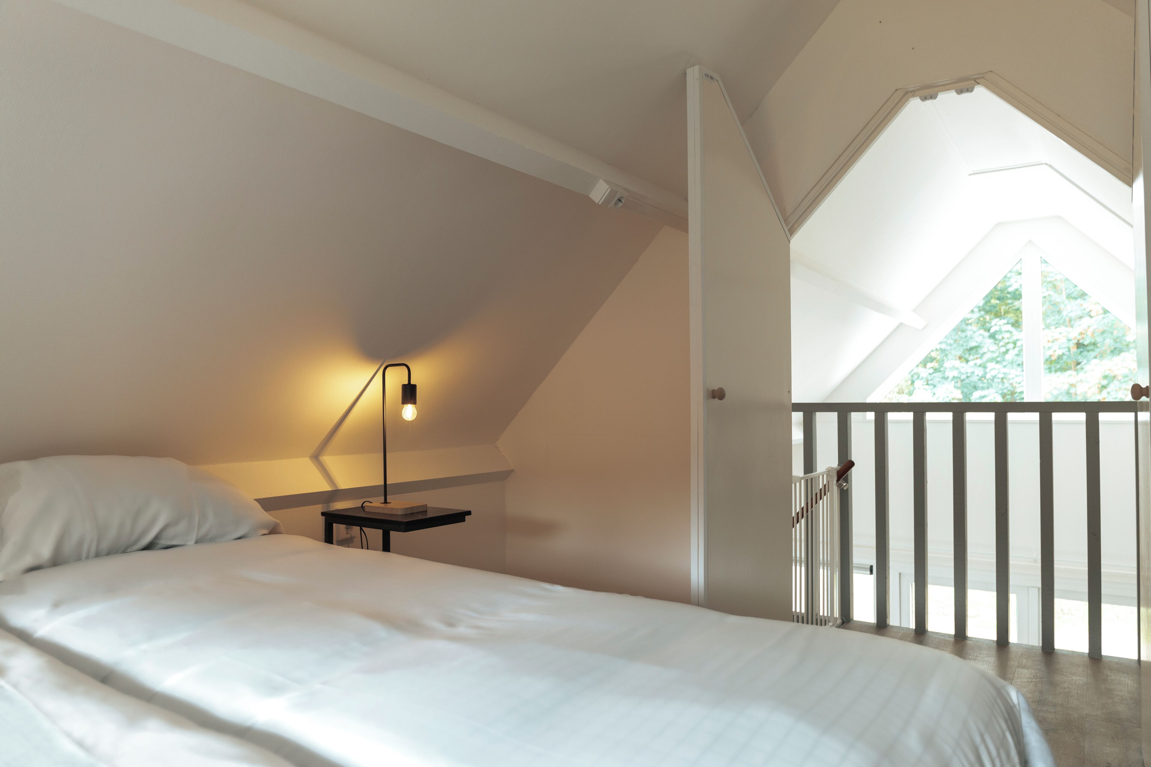 Een slaapkamer met een bed, een nachtkastje, een wandlamp, een raam met uitzicht op groen en een balkon met een balustrade.