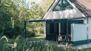 A house with a covered patio, a dining area with chairs, and a large window. - The bungalow Palthe Comfort offers comfort and tranquility in nature for 4 people, with modern amenities and a lovely terrace. (Nieuw Heeten)