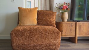 A brown textured sofa with a cushion, positioned in front of a wooden cabinet with a vase of flowers and a television on top. - The Palthe Premium Living bungalow has been recently fully renovated and features a stylish, modern interior. It offers all the comfort for 4 people, surrounded by nature, with a lovely terrace to relax outdoors. (Nieuw Heeten)