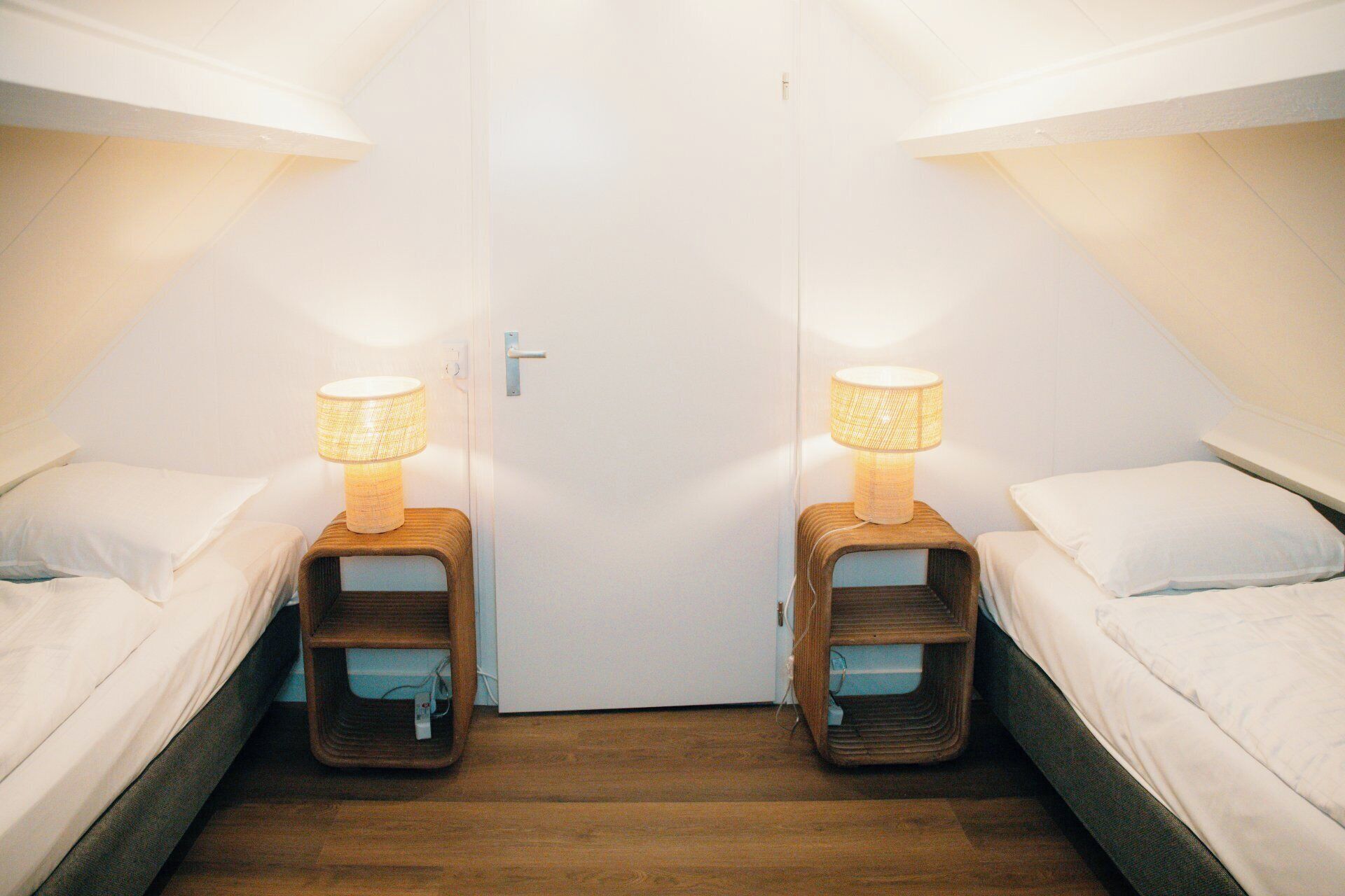 A room with two beds, each with a wooden nightstand and a lamp. There is a white door in the center.