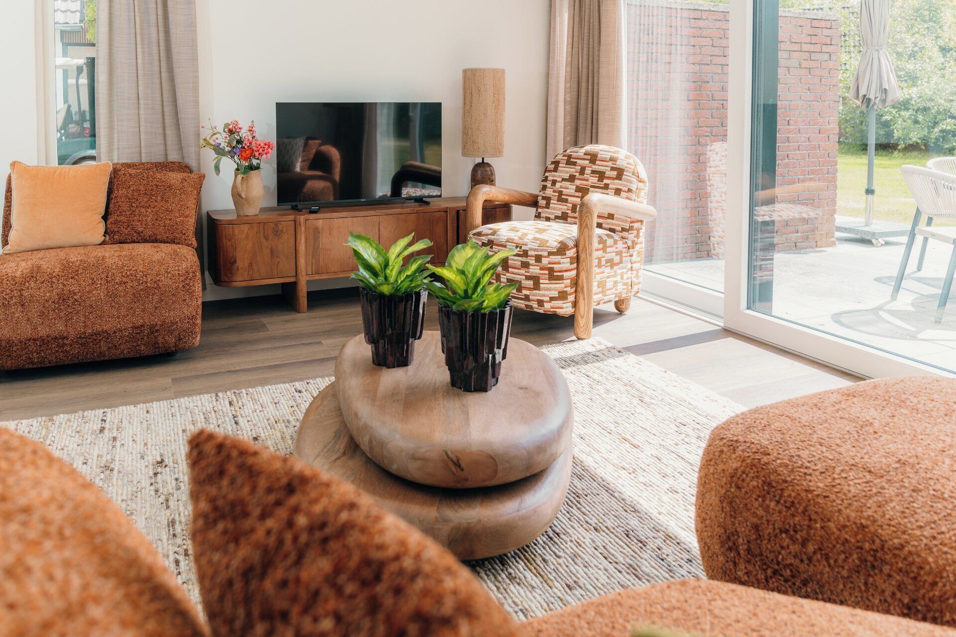 A modern living room with a sofa, armchair, and ottoman, a television, and two potted plants.