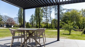 An outdoor patio with a table and chairs, surrounded by a covered area and a view of trees and a residential area. - The Palthe Premium bungalow has been recently fully renovated and features a stylish, modern interior. It offers all the comfort for 4 people, surrounded by nature, with a lovely terrace to relax outdoors. (Nieuw Heeten)
