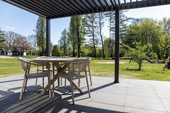 An outdoor patio with a table and chairs, surrounded by a covered area and a view of trees and a residential area.