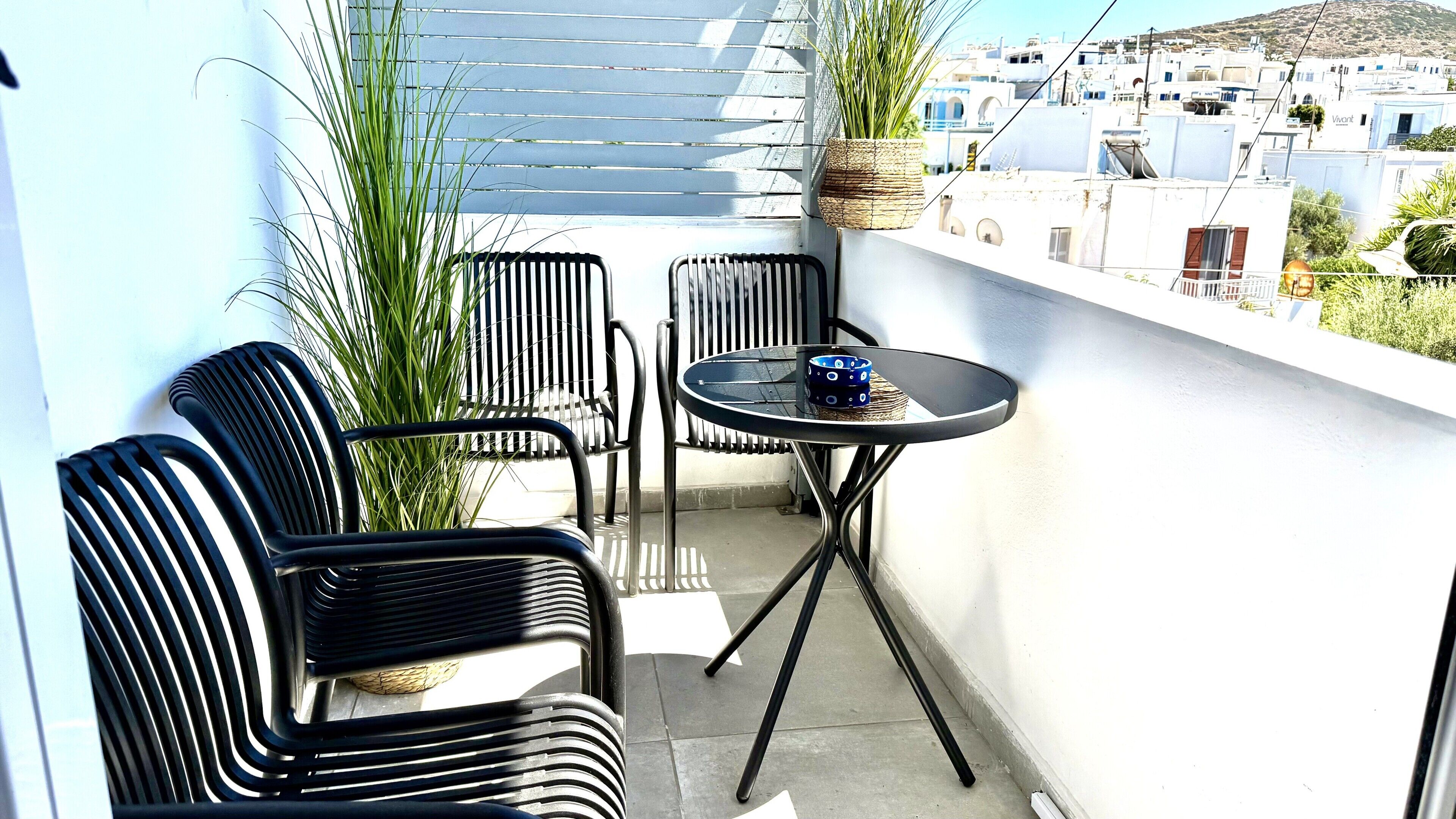 A balcony with two black metal chairs and a small round table, a potted plant, and a view of a white building with blue accents.
