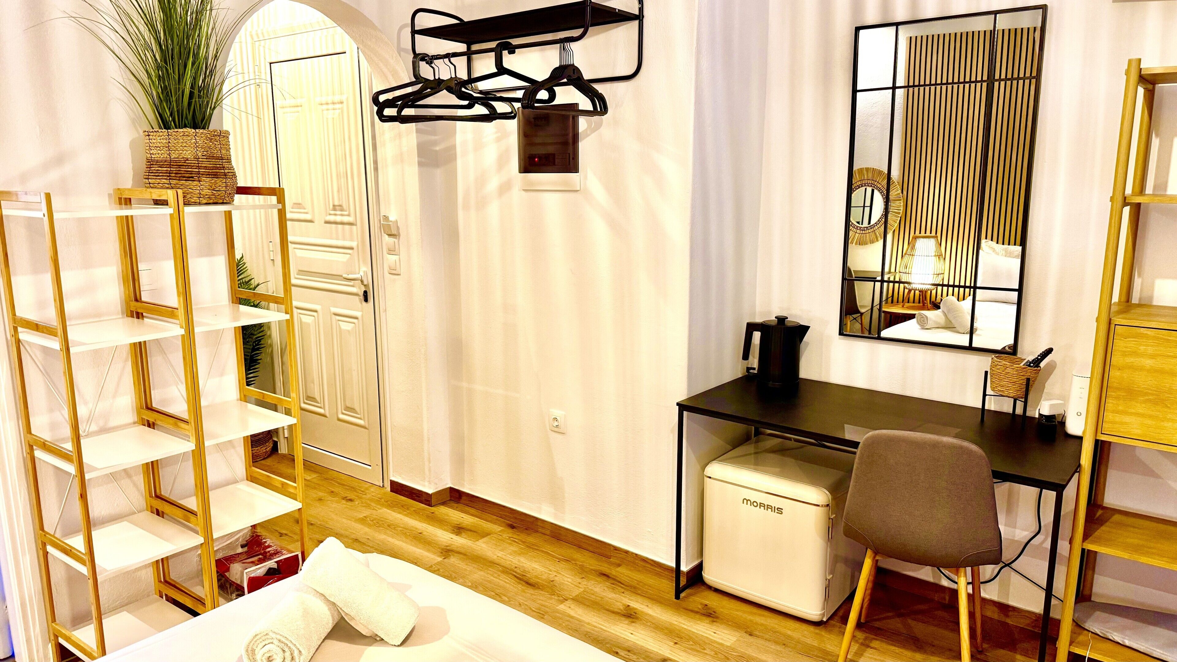 A hotel room with a desk, a chair, a small refrigerator, a hanging coat rack, and a potted plant.
