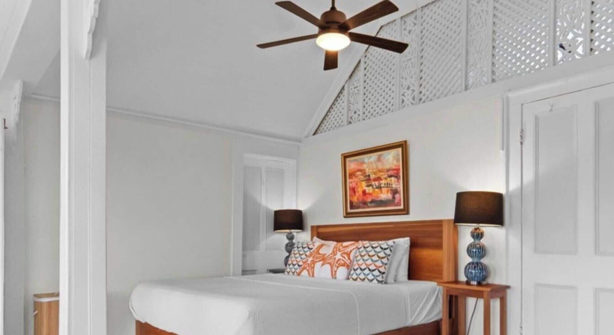 Une chambre à coucher avec un lit, un ventilateur de plafond, un tableau, deux lampes de chevet et une porte.