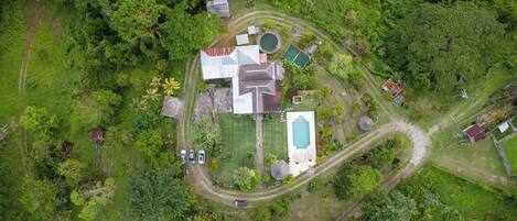 Vue aérienne d’une propriété résidentielle dotée d’une piscine, de courts de tennis et d’une grande maison dont le toit se distingue particulièrement.